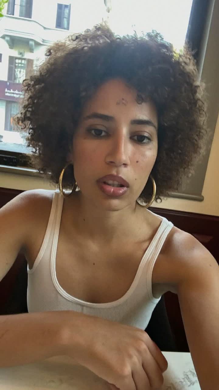 Layla, 42-year-old Lebanese woman, short curly afro, wearing a white ribbed tank top and gold hoops